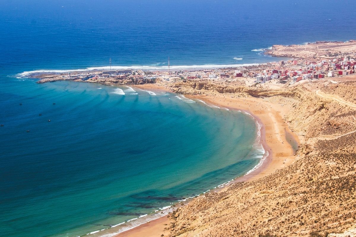 Aerial view of Imsouane Bay in Morocco, with turquoise waves curling toward the sandy shoreline and a coastal village in the distance.
