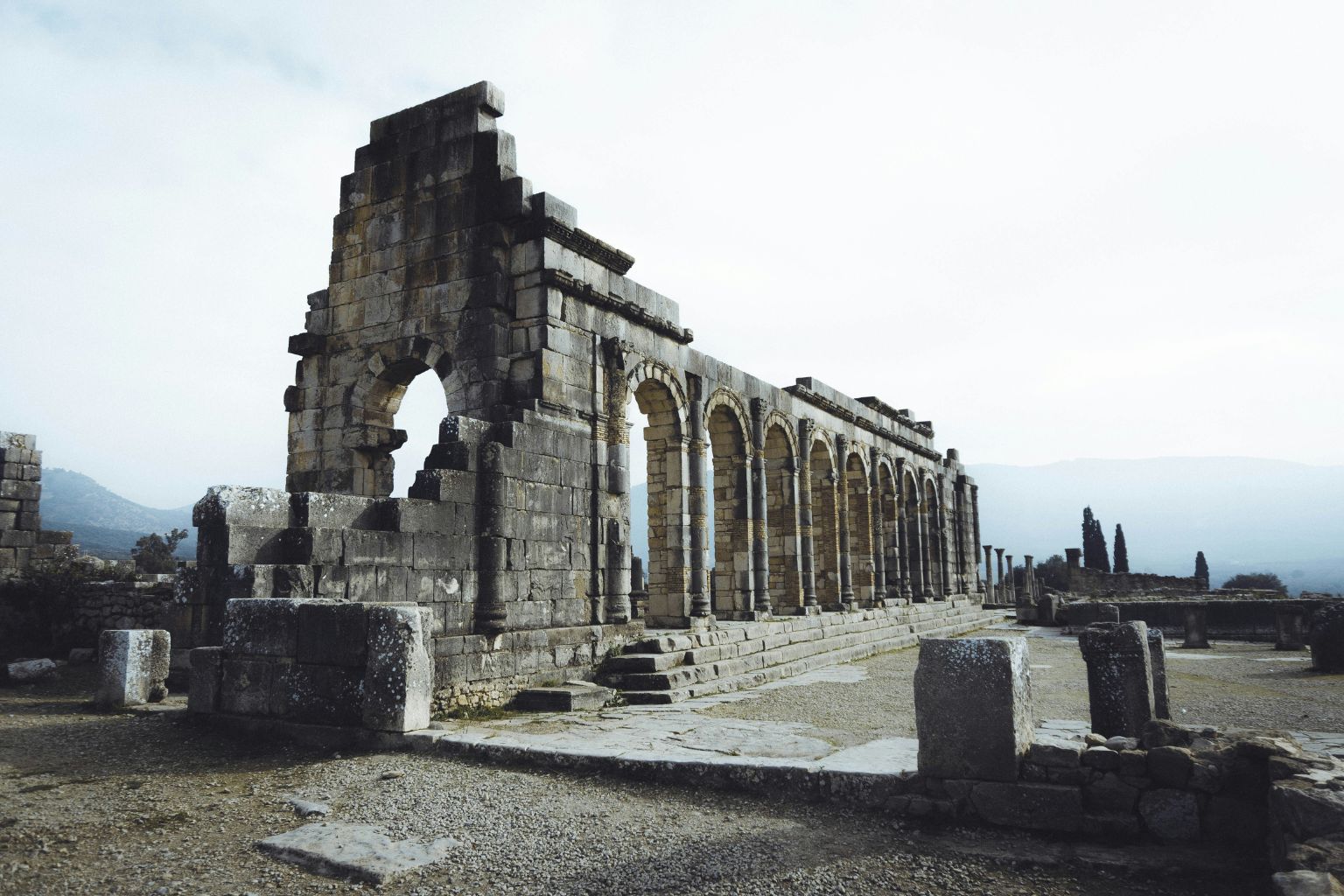 Ruins of the ancient city of Volubilis in Morocco, showing Roman arches and stone structures against a mountainous backdrop.