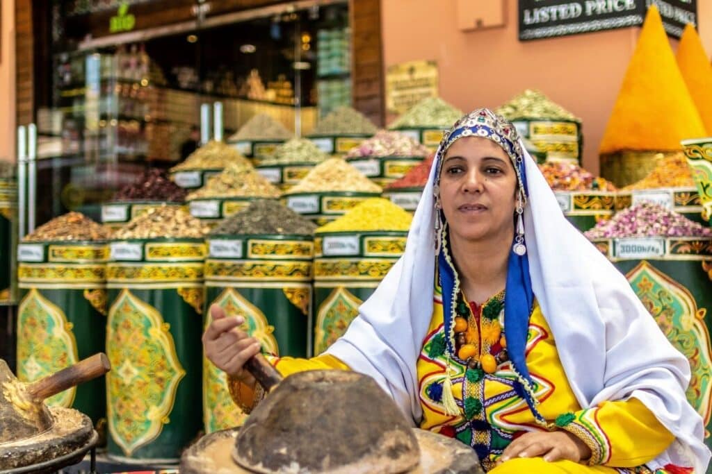 A Moroccan Amazigh woman in traditional dress grinding spices by hand in front of a colorful Moroccan spices shop.