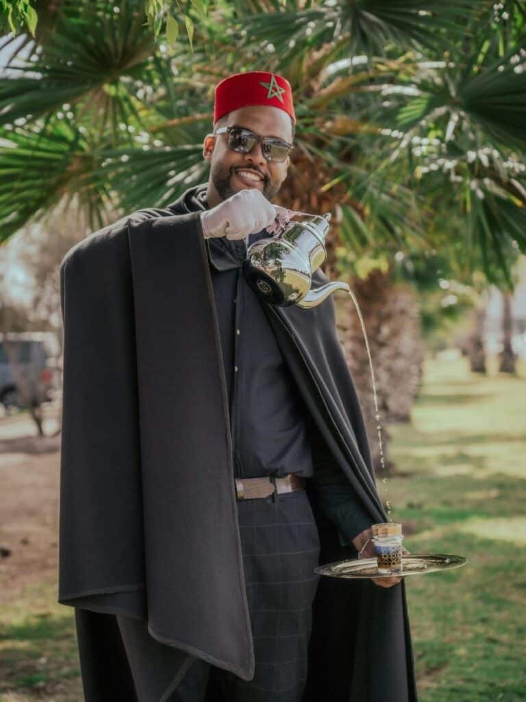 Man in traditional Moroccan attire pouring mint tea from a silver teapot held high above a decorated glass on a tray.