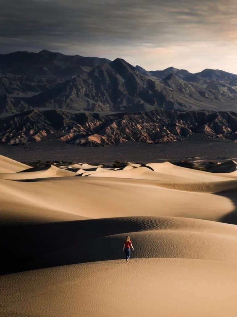 A person walks across the golden sand dunes of Zagora with rugged mountains rising in the background under a moody sky.